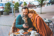 Cute middle-aged lovers are having breakfast in restaurant outside. They are sitting and holding hands. Man is using smartphone and smiling. Woman is laying head on his shoulder